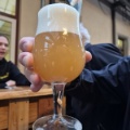 Hand, die in einer Bar ein bauchiges Glas mit trübem, hellorangem Bier und dichter Schaumkrone in die Kamera hält